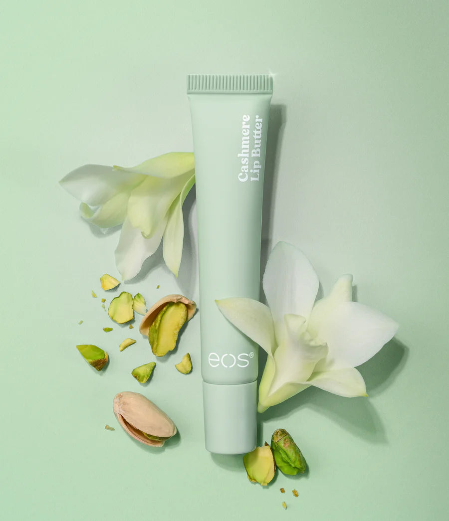 A pale mint green tube of EOS Cashmere Lip Butter is styled on a matching mint green background with white orchid flowers and pistachios around it. The vertical text reads 'Cashmere Lip Butter' and the brand 'eos' is visible near the bottom. Purchase from www.americanbodycare.co.uk
