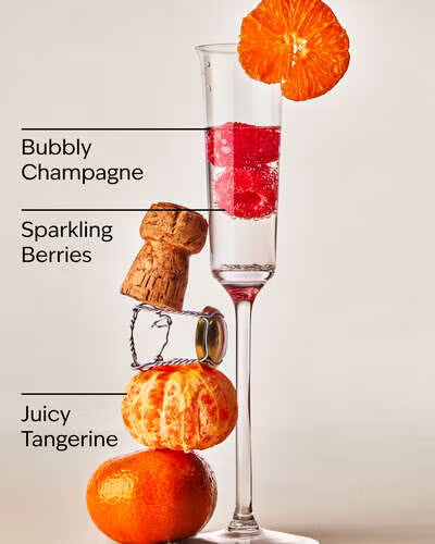 Champagne flute with red berries, cork, and tangerines on a light background. Car fragrance refill labeled 'Champagne Toast' by White Barn on a beige background. Purchase from www.americanbodycare.co.uk 