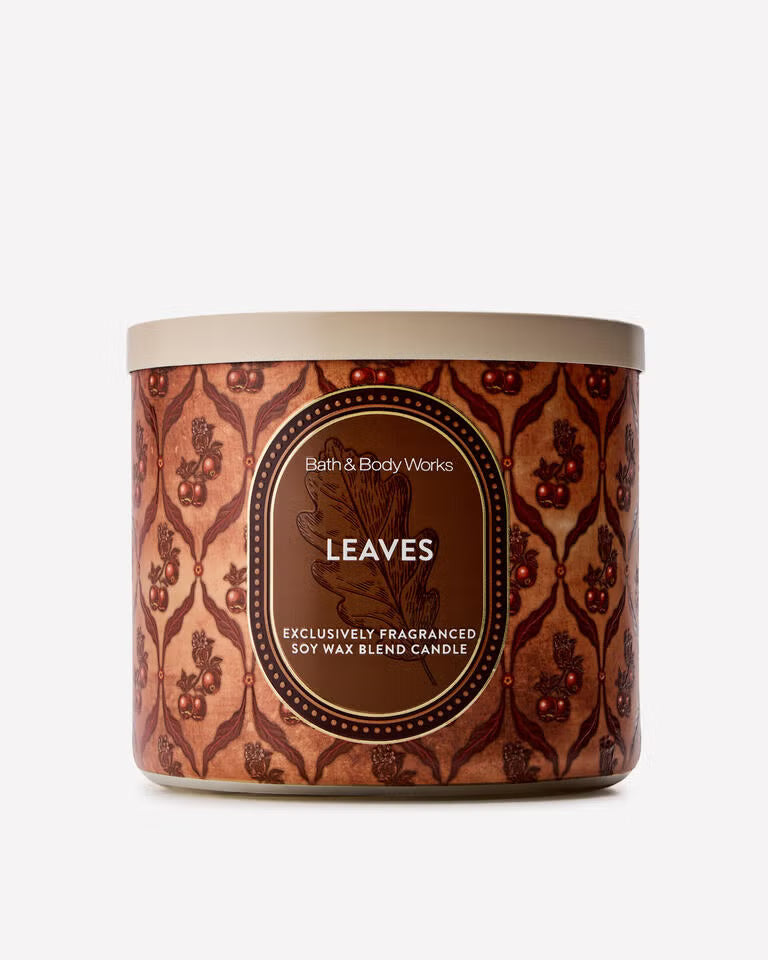 A Bath & Body Works 'Leaves' soy wax blend candle in a cylindrical tin with a brown and copper patterned label and a beige lid, set against a plain light gray background. Purchase from www.americanbodycare.co.uk 