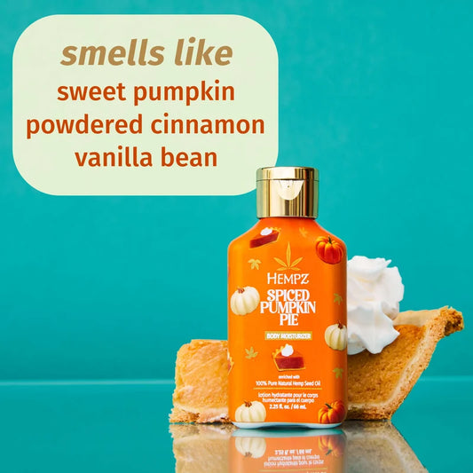 A bottle of Hempz Spiced Pumpkin Pie body moisturizer stands on a reflective teal surface with autumn-themed props: a slice of pumpkin pie, whipped cream, and small pumpkins. A large text bubble reads 'smells like sweet pumpkin powdered cinnamon vanilla bean. purchase from www.americanbodycare.co.uk