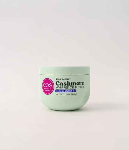A light mint green jar of EOS Cashmere Whipped Oil Butter with a pink circular logo, centered on a light gray background. Purchase from www.americanbodycare.co.uk 