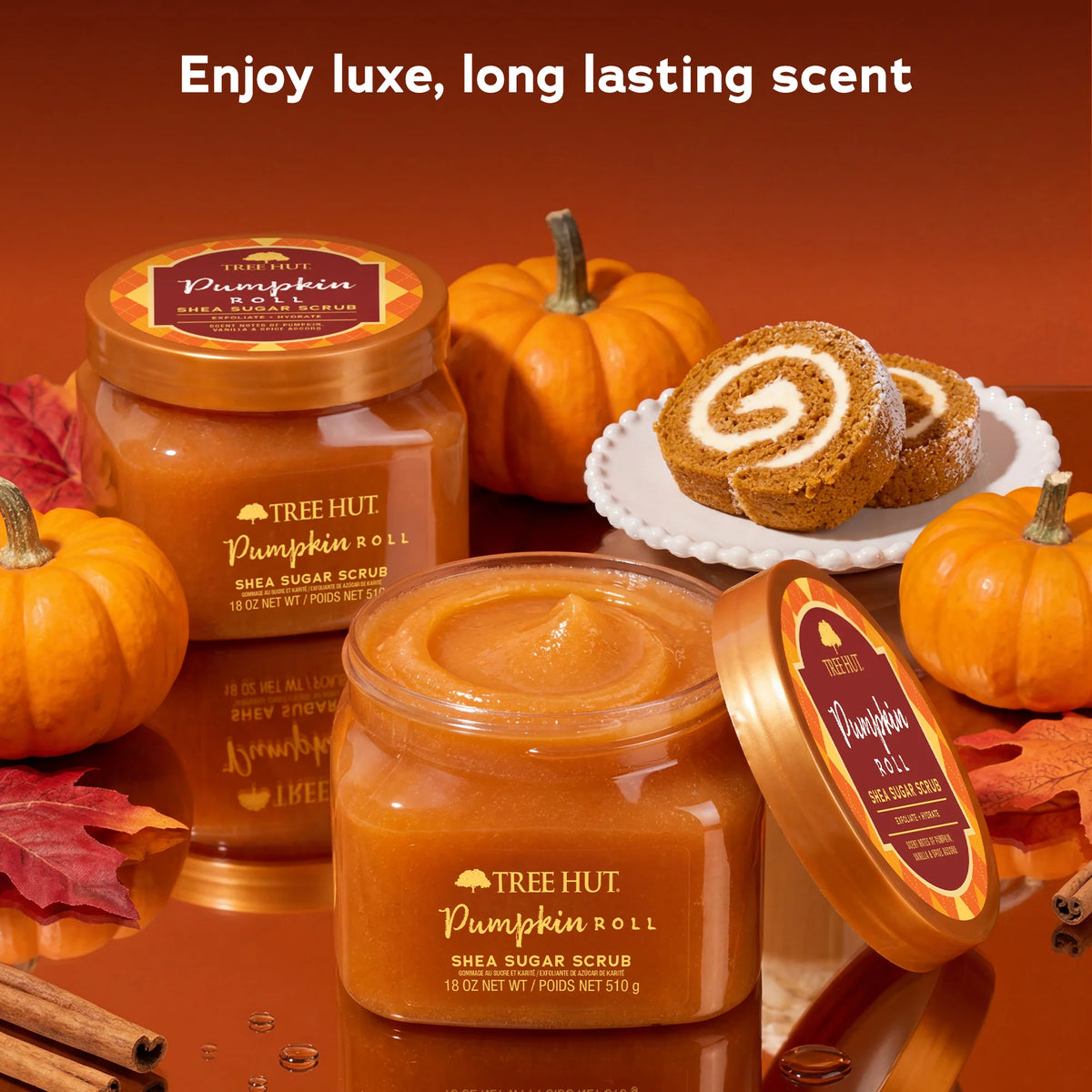 Three jars of Tree Hut Pumpkin Roll Shea Sugar Scrub are arranged on a warm brown surface with decorative pumpkins, fall leaves, cinnamon sticks, and a slice of pumpkin roll cake. White text at the top reads 'Enjoy luxe, long lasting scent'. www.americanbodycare.co.uk
