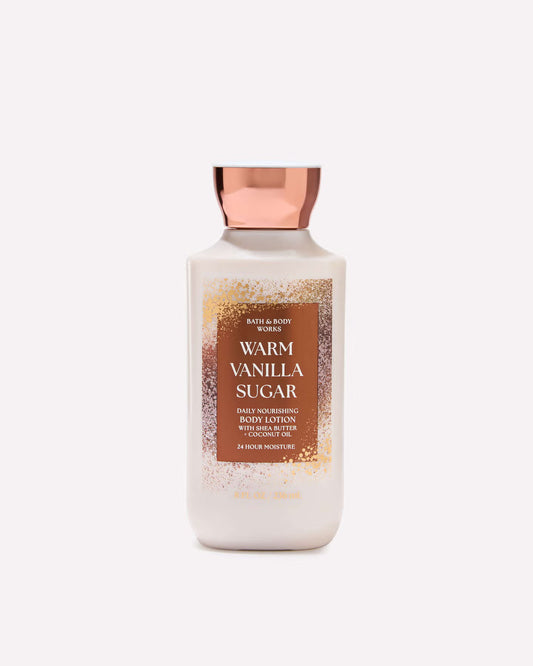 A bottle of Bath & Body Works Warm Vanilla Sugar body moisturizing body lotion, featuring a white bottle with a metallic rose gold cap and a brown label with gold glitter details, set against a solid off-white background. purchase from www.americanbodylotion.co.uk