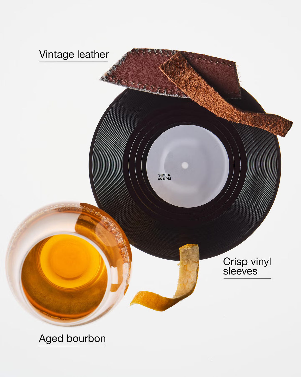 A styled flat-lay on a white background featuring a black vinyl record, a slice of vintage brown leather, a curled piece of aged brown leather, and a glass of amber bourbon with a gold rim. Text labels read 'Vintage leather', 'Crisp vinyl sleeves', and 'Aged bourbon'. Bath & Body Works Bourbon Oak Records soy wax blend candle in a round glass jar with a black lid and a warm brown label decorated with vinyl records and autumn leaves. purchase from www.americanbodycare.co.uk