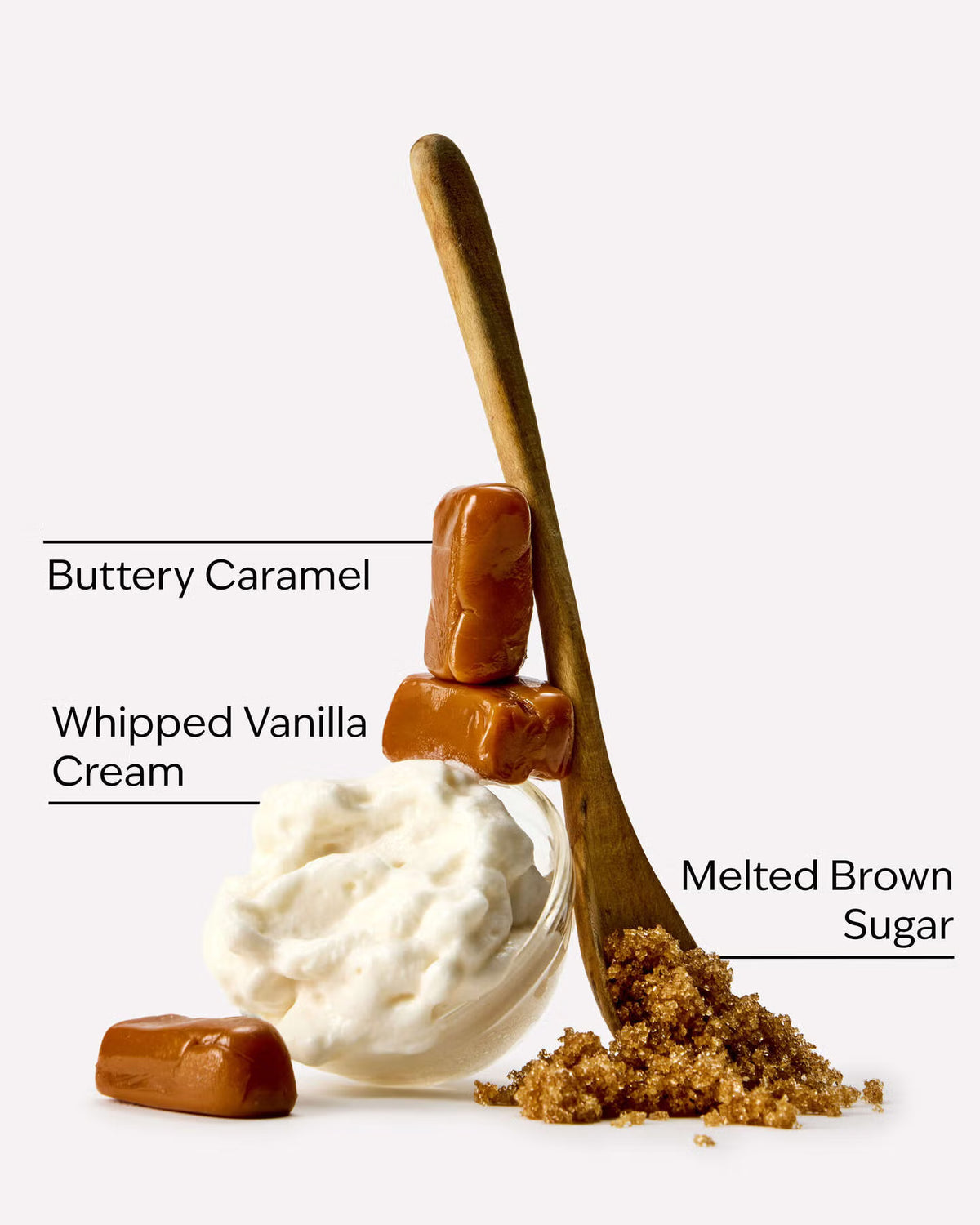 A styled food product shot on a light gray background showing a small glass bowl of whipped vanilla cream, two buttery caramel candies, a wooden spoon filled with melted brown sugar, and text labels identifying each ingredient. A bottle of Bath & Body Works Caramel Vanilla body wash, featuring a warm amber-brown liquid inside a clear bottle with a metallic gold cap and a cream-colored label, set against a solid light gray background. purchase from www.americanbodycare.co.uk