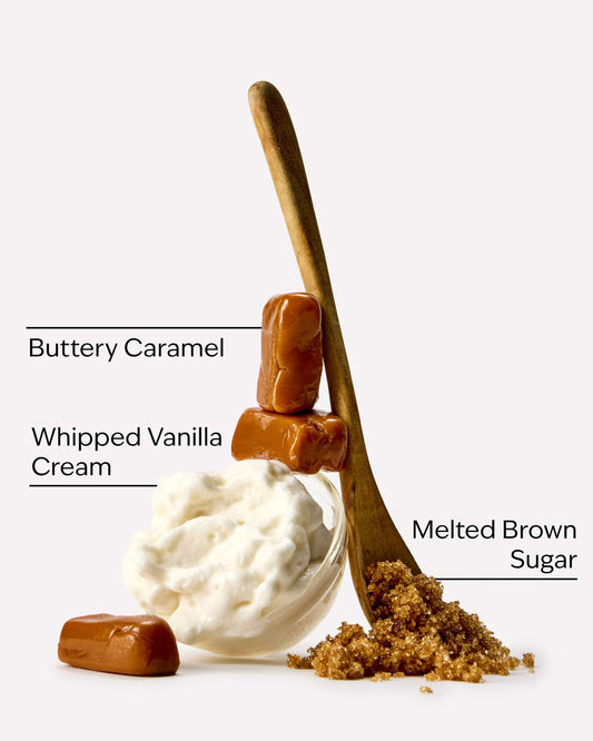 A styled food product shot on a light gray background showing a small glass bowl of whipped vanilla cream, two buttery caramel candies, a wooden spoon filled with melted brown sugar, and text labels identifying each ingredient. A bottle of Bath & Body Works Caramel Vanilla body wash, featuring a warm amber-brown liquid inside a clear bottle with a metallic gold cap and a cream-colored label, set against a solid light gray background. purchase from www.americanbodycare.co.uk