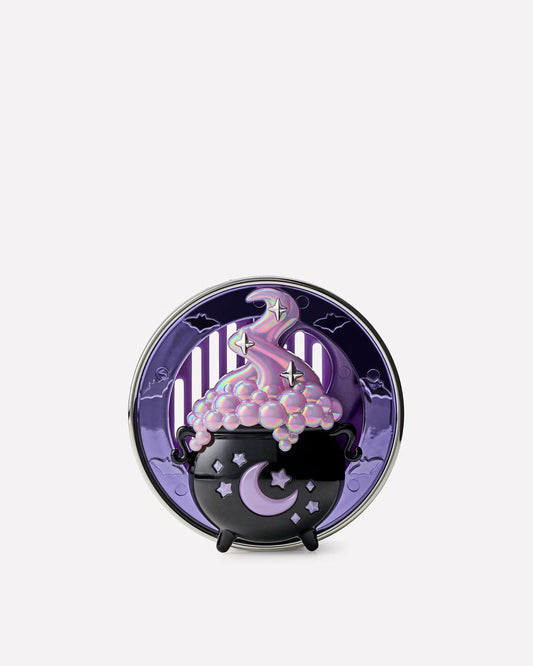 A circular decorative item featuring a purple and black witch-themed design with a cauldron, moon, stars, and pink glittery substance spilling over, displayed on a small black stand against a light gray background. purchase from www.americanbodycare.co.uk