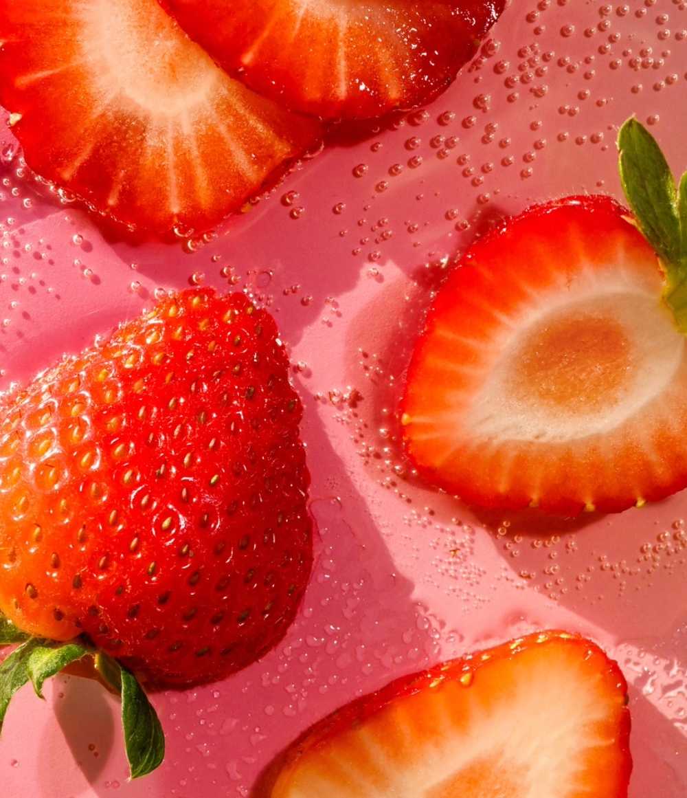 Strawberries, both whole and sliced, on a pink surface with water droplets. Purchase from americanbodycare.co.uk