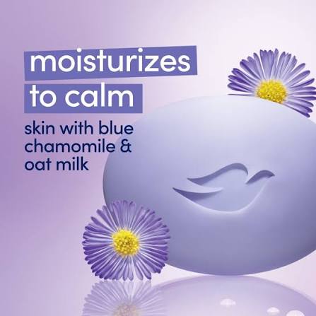 A lavender-colored bar of soap with a peace sign embossed on it, decorated with two purple chamomile flowers with yellow centers. The background is a soft lavender gradient with text that reads 'moisturizes to calm skin with blue chamomile & oat milk'. Dove Anti-Stress Moisturizing Cream Beauty Bar packaging, featuring a blue oval bar soap graphic on a white and blue box with a gold Dove logo. Purchase from www.americanbodycare.co.uk 