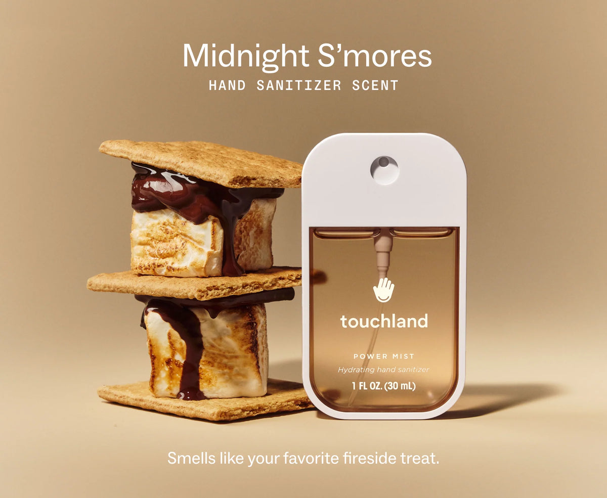 A Touchland hand sanitizer spray bottle labeled 'Midnight S'mores' stands beside a stacked s'more with toasted bread, marshmallow, and chocolate drizzle on a warm beige background. Text reads 'Midnight S'mores HAND SANITIZER SCENT' and 'Smells like your favorite fireside treat. Purchase from www.americanbodycare.co.uk